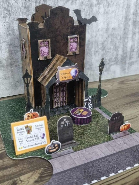 3Quarter Designs Witchwood Charm Tarot Store miniature building front view showing dark gothic facade with damask wallpaper panels, crooked roofline with bat die-cuts, Witchwood Charm sign with crystal ball, arched porch entrance, street lamp die-cuts, wrought iron picket fence, RIP tombstone, jack-o-lantern, black cat, tarot card services menu sign, and purple striped crystal ball table on grass graveyard base board