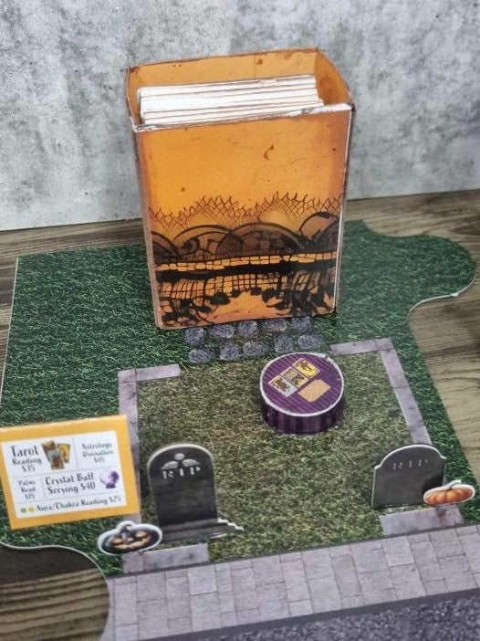 3Quarter Designs Witchwood Charm Tarot Store hidden star book sitting on the grass graveyard base board showing the orange Halloween gothic cover with spider web and landscape scene, RIP tombstones, purple crystal ball table with tarot card lid, and tarot card services menu sign