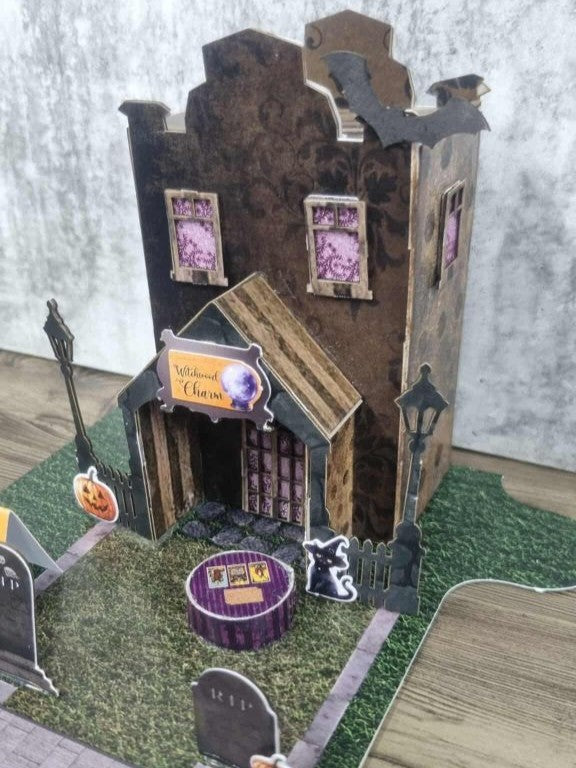 3Quarter Designs Witchwood Charm Tarot Store miniature building angled front view showing dark gothic two-storey facade, bat die-cuts on roofline, Witchwood Charm crystal ball sign, arched porch with striped timber roof, purple lace upper windows, street lamp, wrought iron fence, black cat die-cut, purple striped crystal ball table, and cobblestone road section on jigsaw base board