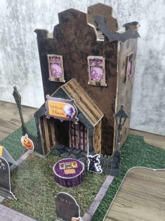 3Quarter Designs Witchwood Charm Tarot Store miniature building angled front view showing dark gothic two-storey facade, bat die-cuts on roofline, Witchwood Charm crystal ball sign, arched porch with striped timber roof, purple lace upper windows, street lamp, wrought iron fence, black cat die-cut, purple striped crystal ball table, and cobblestone road section on jigsaw base board