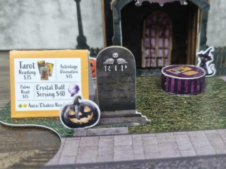 Close-up of 3Quarter Designs Witchwood Charm Tarot Store miniature building showing tarot card services menu sign listing Tarot Reading, Astrology Divination, Palms Read, Crystal Ball Scrying and Aura Chakra Reading, RIP tombstone with skull detail, jack-o-lantern die-cut, purple striped crystal ball table with tarot cards, black cat die-cut, and arched lace panel door in background