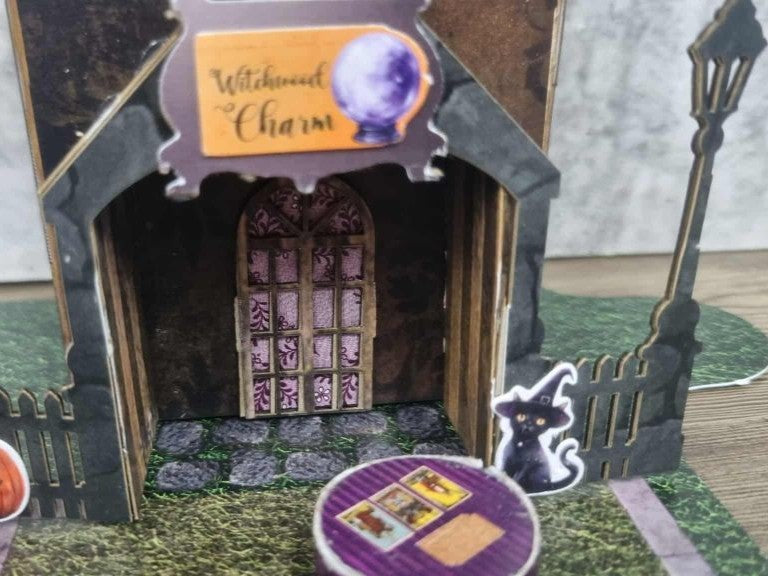  Close-up of 3Quarter Designs Witchwood Charm Tarot Store miniature building porch showing Witchwood Charm sign with glowing crystal ball, arched doorway with ornate floral lace panel double doors, cobblestone porch floor, black cat die-cut, purple tarot card table, and wrought iron street lamp on grass base board