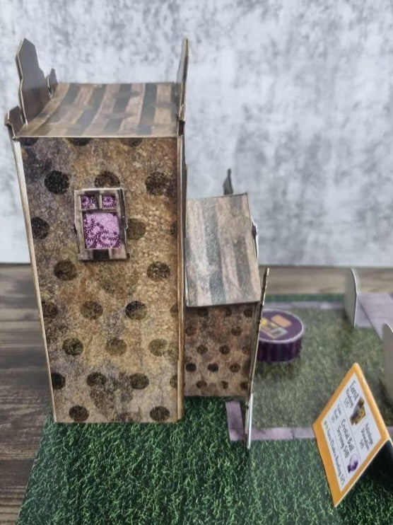 3Quarter Designs Witchwood Charm Tarot Store miniature building left side view showing dark textured spotty gothic paper walls, striped timber porch roof, purple lace framed upper window, purple striped table, tarot card services menu sign, and grass jigsaw base board