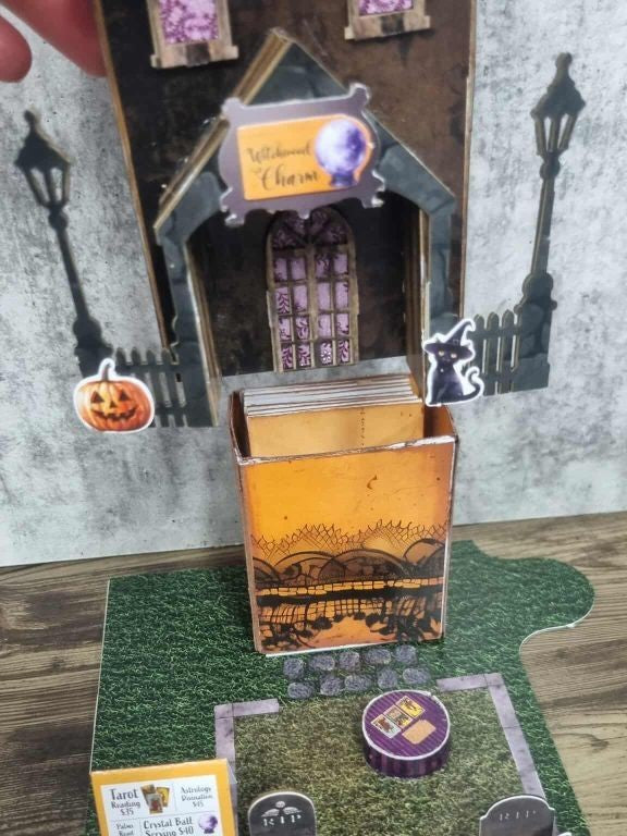 3Quarter Designs Witchwood Charm Tarot Store miniature building being lifted to reveal a hidden orange Halloween star book underneath, showing the Witchwood Charm sign, arched lace panel door, jack-o-lantern, black cat, street lamp die-cuts, tarot card services menu sign, RIP tombstones, and purple crystal ball table on grass graveyard base board