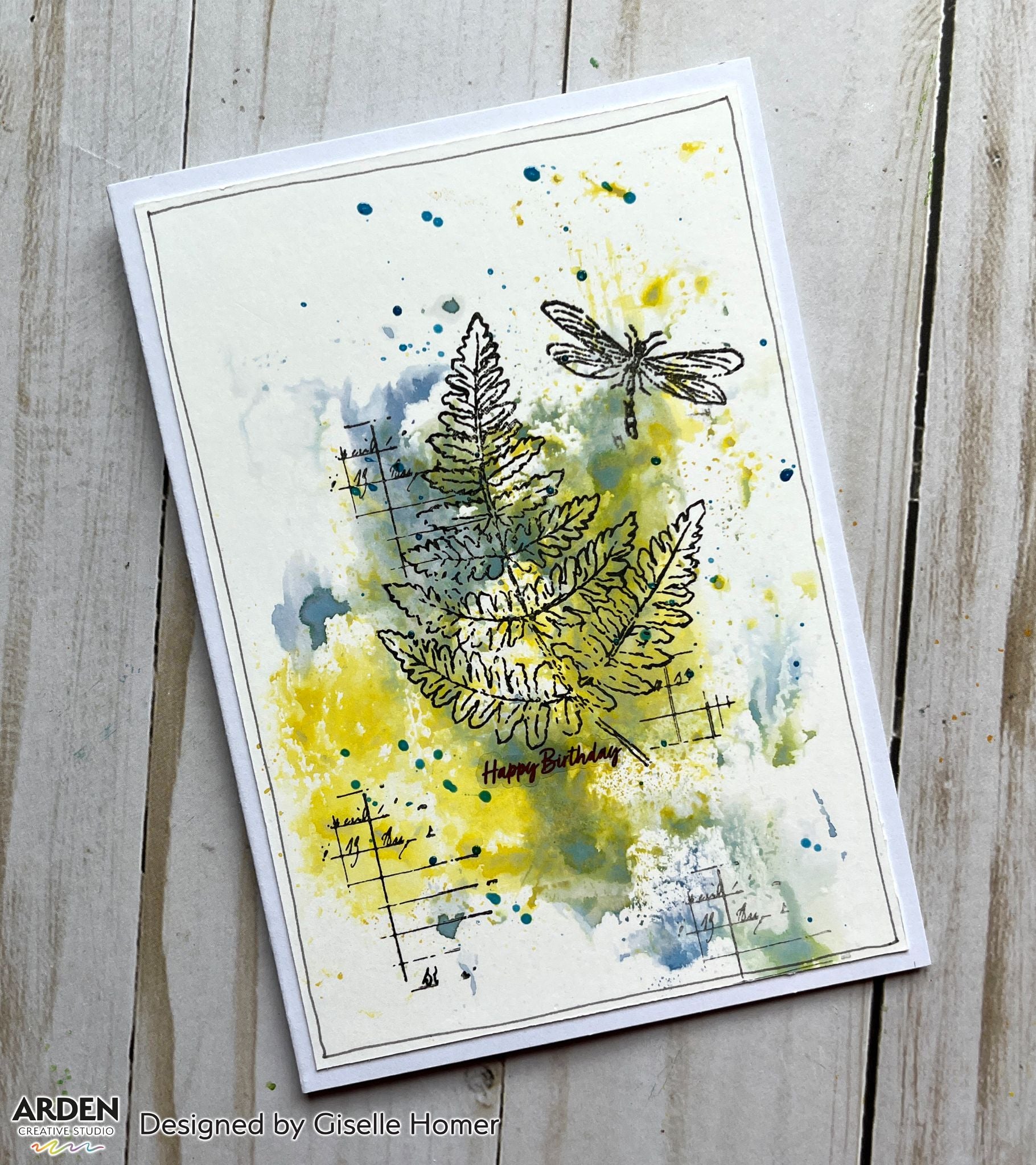 Handmade birthday card sample using Arden Creative Studio Hidden Creek collection, featuring fern and botanical leaf stamping in yellow-green watercolour tones, dragonfly stamp, grid background stamping, ink splatters, and a "Happy Birthday" sentiment.