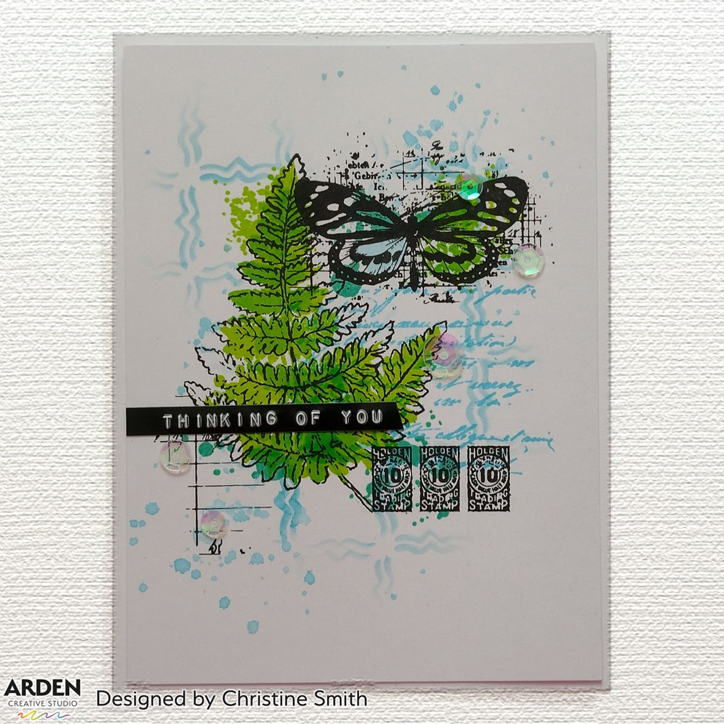 Handmade card sample using Arden Creative Studio Hidden Creek collection, featuring bold green fern stamping, butterfly stamp, teal ink splatters, vintage postage stamp embellishments, script background stamping, sequins, and a "Thinking of You" sentiment banner.