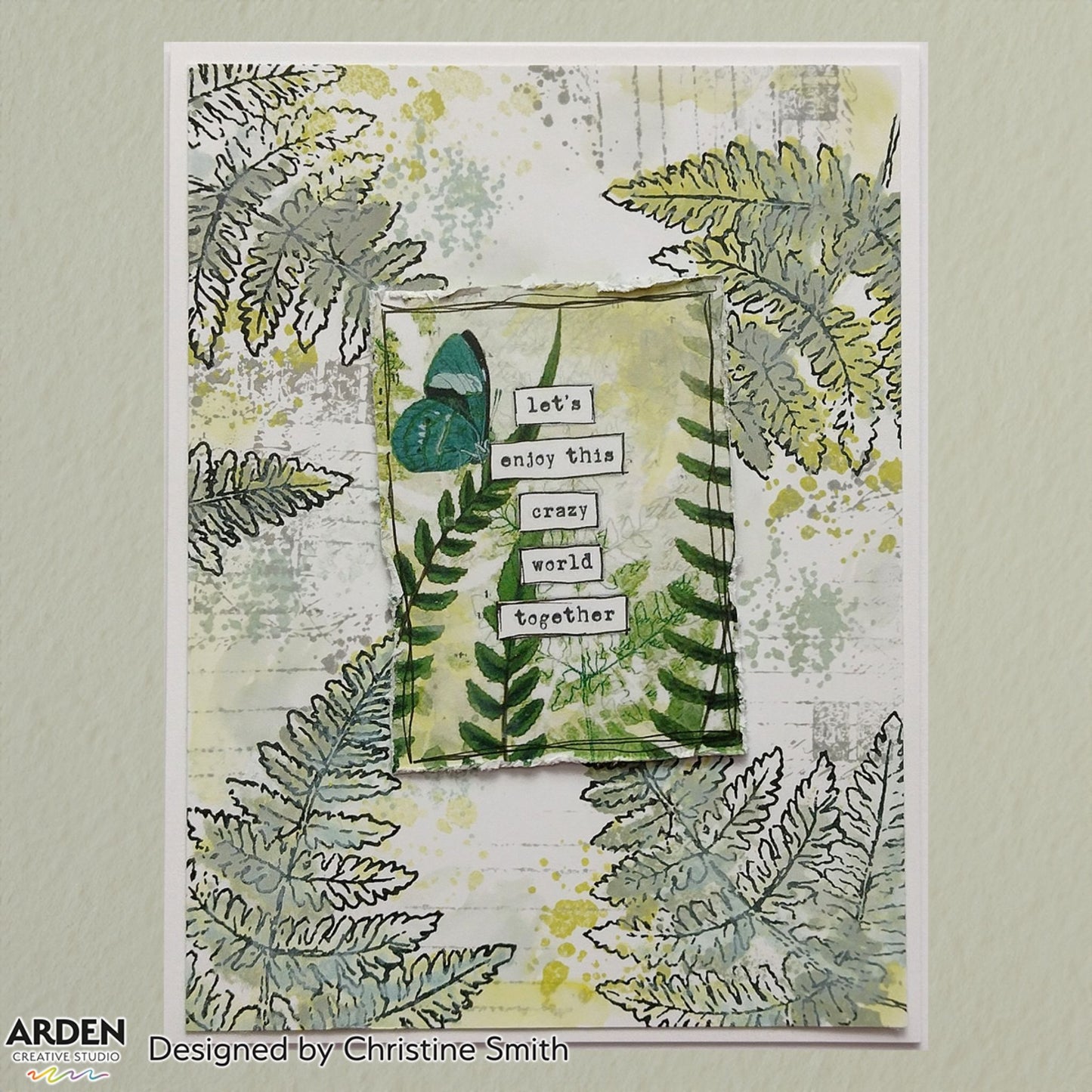 Handmade card sample using Arden Creative Studio Hidden Creek collection, featuring layered fern stamping in green and grey tones, teal butterfly, yellow ink splatters, torn paper focal panel, and a stacked "Let's Enjoy This Crazy World Together" sentiment.