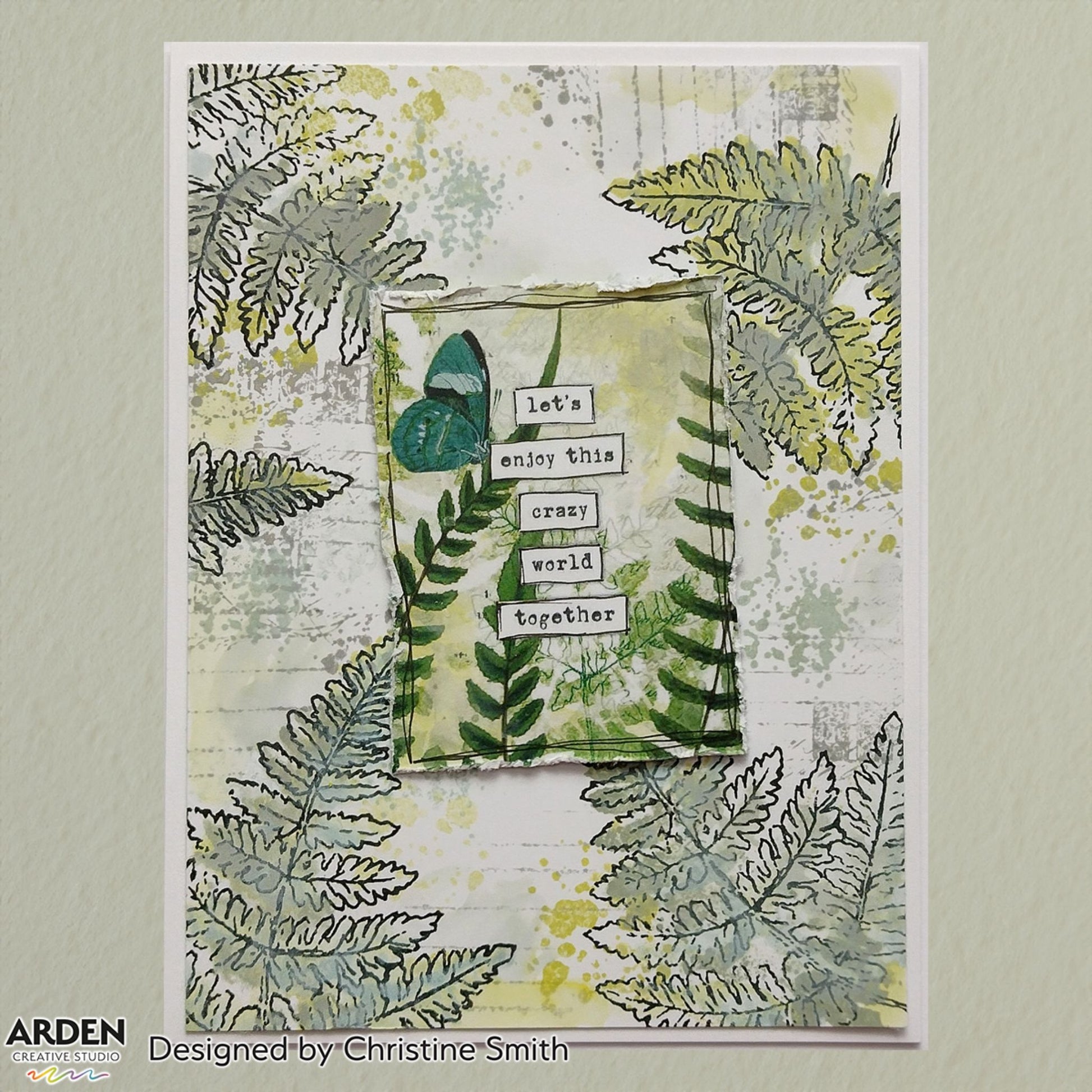Handmade card sample using Arden Creative Studio Hidden Creek collection, featuring layered fern stamping in green and grey tones, teal butterfly, yellow ink splatters, torn paper focal panel, and a stacked "Let's Enjoy This Crazy World Together" sentiment.