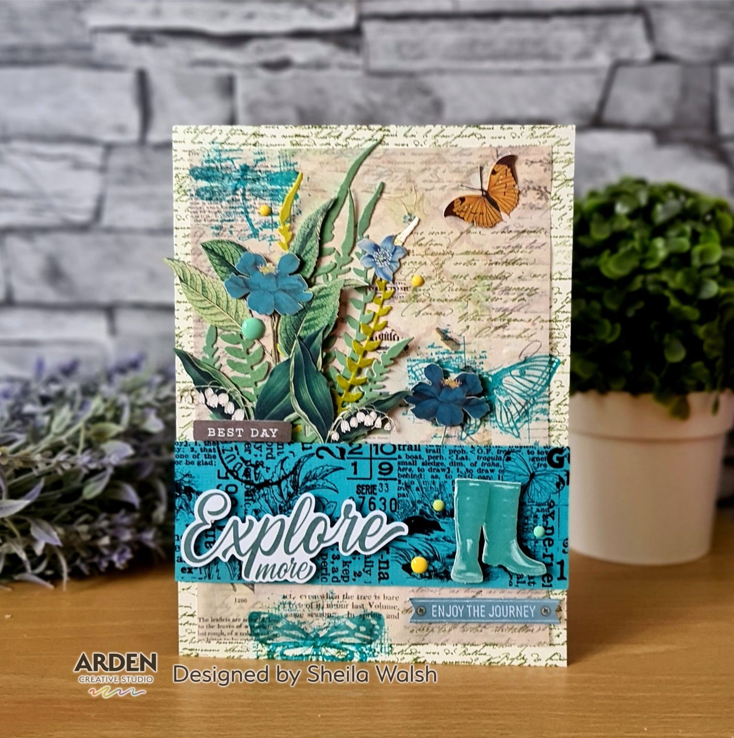 Handmade "Explore More" card sample using Arden Creative Studio Hidden Creek collection, featuring layered botanical die-cuts in teal and green, orange butterfly, wellington boot die-cuts, teal typography background stamping, blue floral embellishments, yellow enamel dots, and "Best Day / Enjoy the Journey" sentiments.