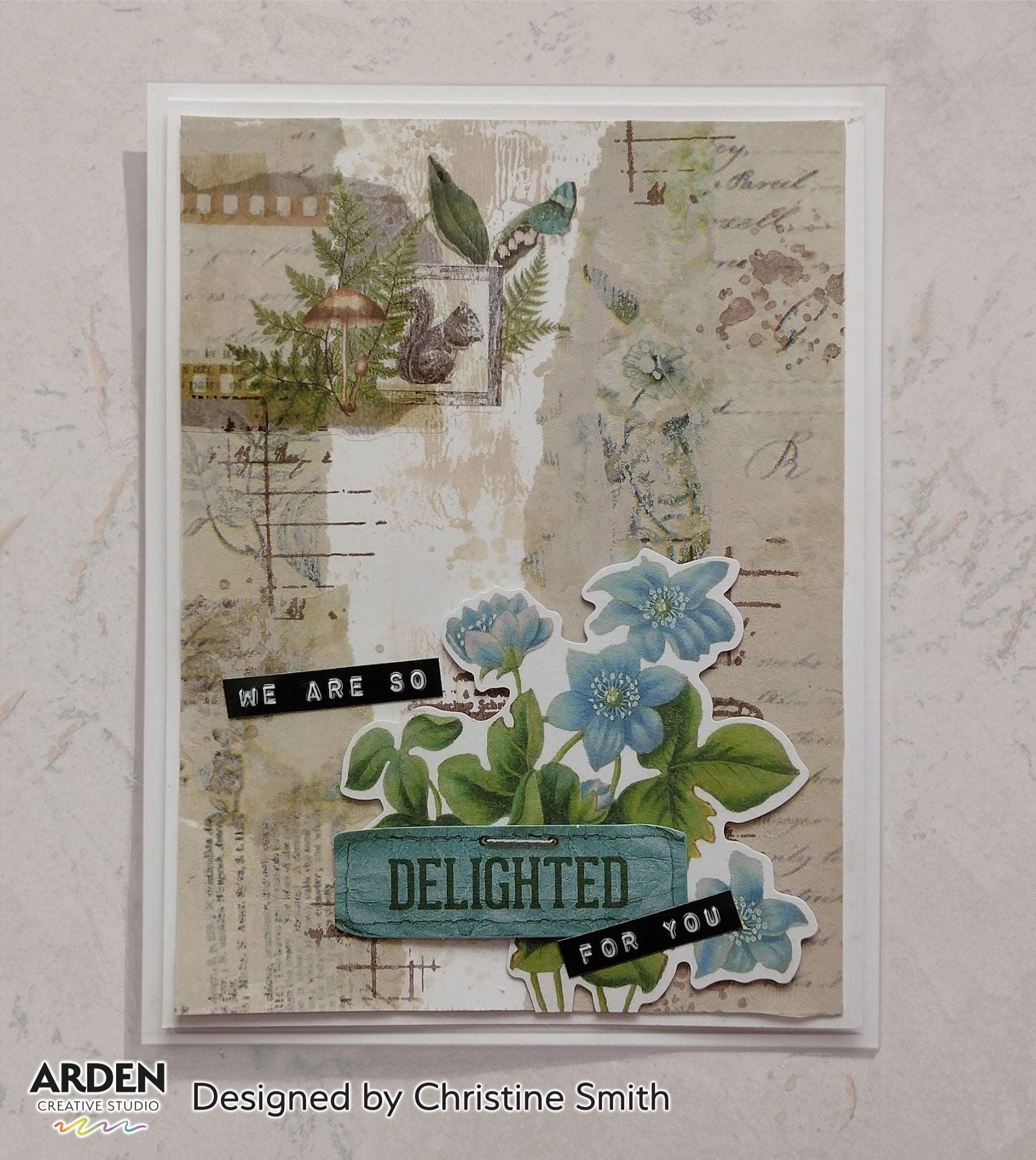 Handmade "We Are So Delighted For You" card sample using Arden Creative Studio Hidden Creek collection, featuring die-cut blue floral focal cluster, squirrel and fern PET Tape panel, mixed media ink blending on a vintage newsprint and script background.