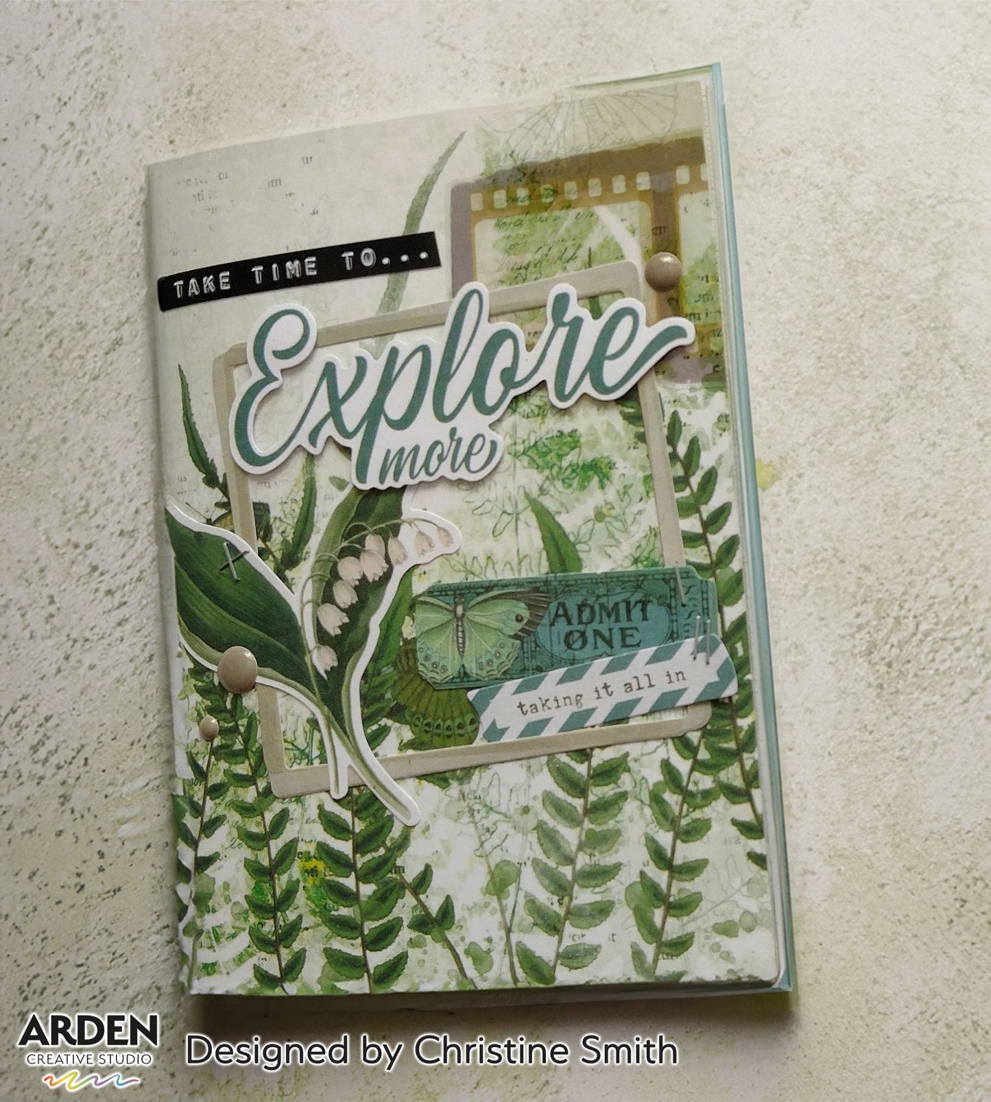 Handmade journal cover sample using Arden Creative Studio Hidden Creek collection, featuring an "Explore More" die-cut title, lily of the valley die-cut, film strip and vintage ticket ephemera on a botanical fern patterned paper background.