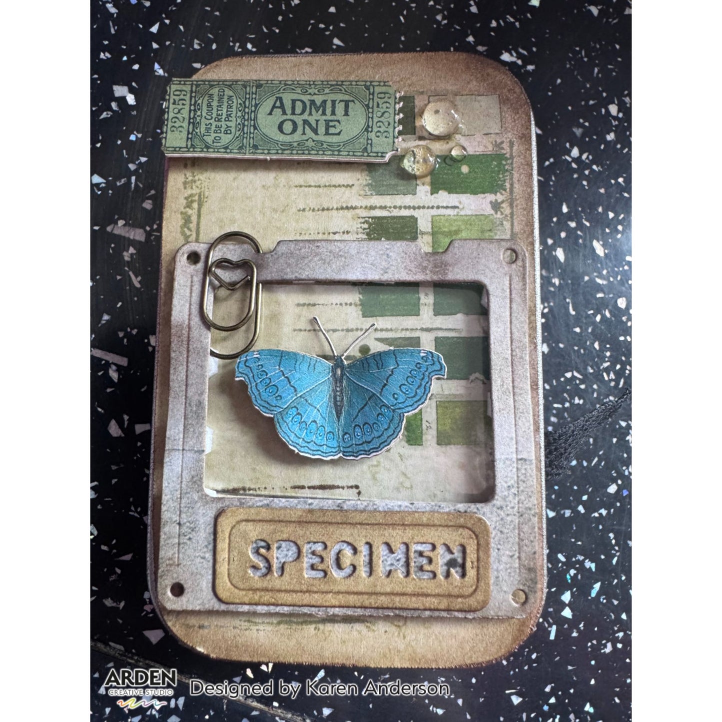 Vintage tin altered art sample using Arden Creative Studio Hidden Creek collection, featuring a teal butterfly die-cut specimen focal point, "Admit One" ticket ephemera, green grid patterned paper, and a heart paper clip embellishment.