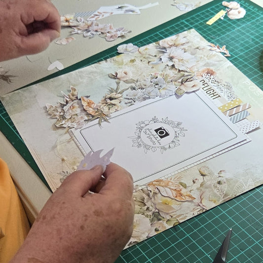 Close-up of hands arranging die-cut floral elements on a scrapbook layout with vintage botanical papers and journaling space