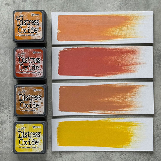 Four Ranger Distress Oxide ink pads from Kit 2 with swatched colours: Spiced Marmalade, Crackling Campfire, Rusty Hinge and Mustard Seed, shown on white paper beside each labelled pad.
