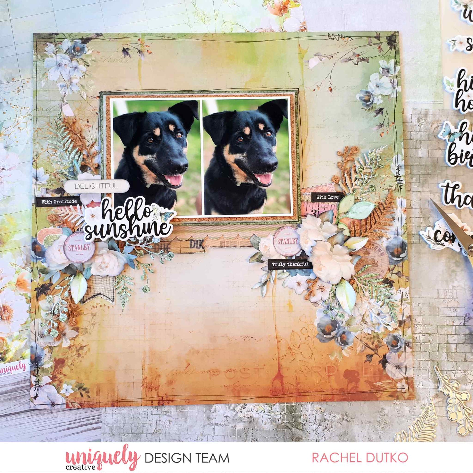 Scrapbook layout titled "Hello Sunshine" featuring two photos of a black dog in a decorative frame, surrounded by cream and blue floral die-cuts and fern embellishments on warm-toned distressed patterned paper, made with Uniquely Creative Delighted Curiosities