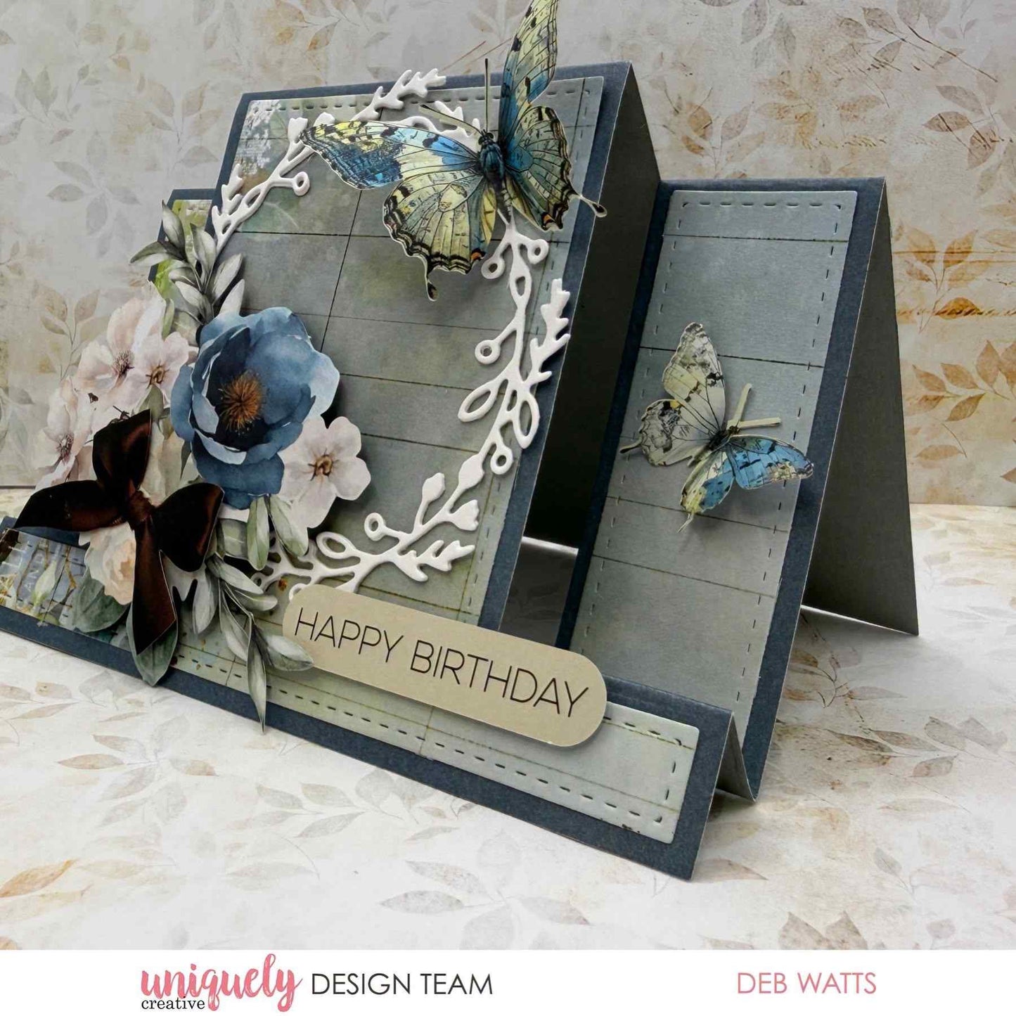Handmade easel birthday card sample using the Uniquely Creative Leafy Wreath Die, featuring a white die-cut wreath framing a blue floral arrangement with paper butterflies on a slate blue stitched panel, with a "Happy Birthday" sentiment 
