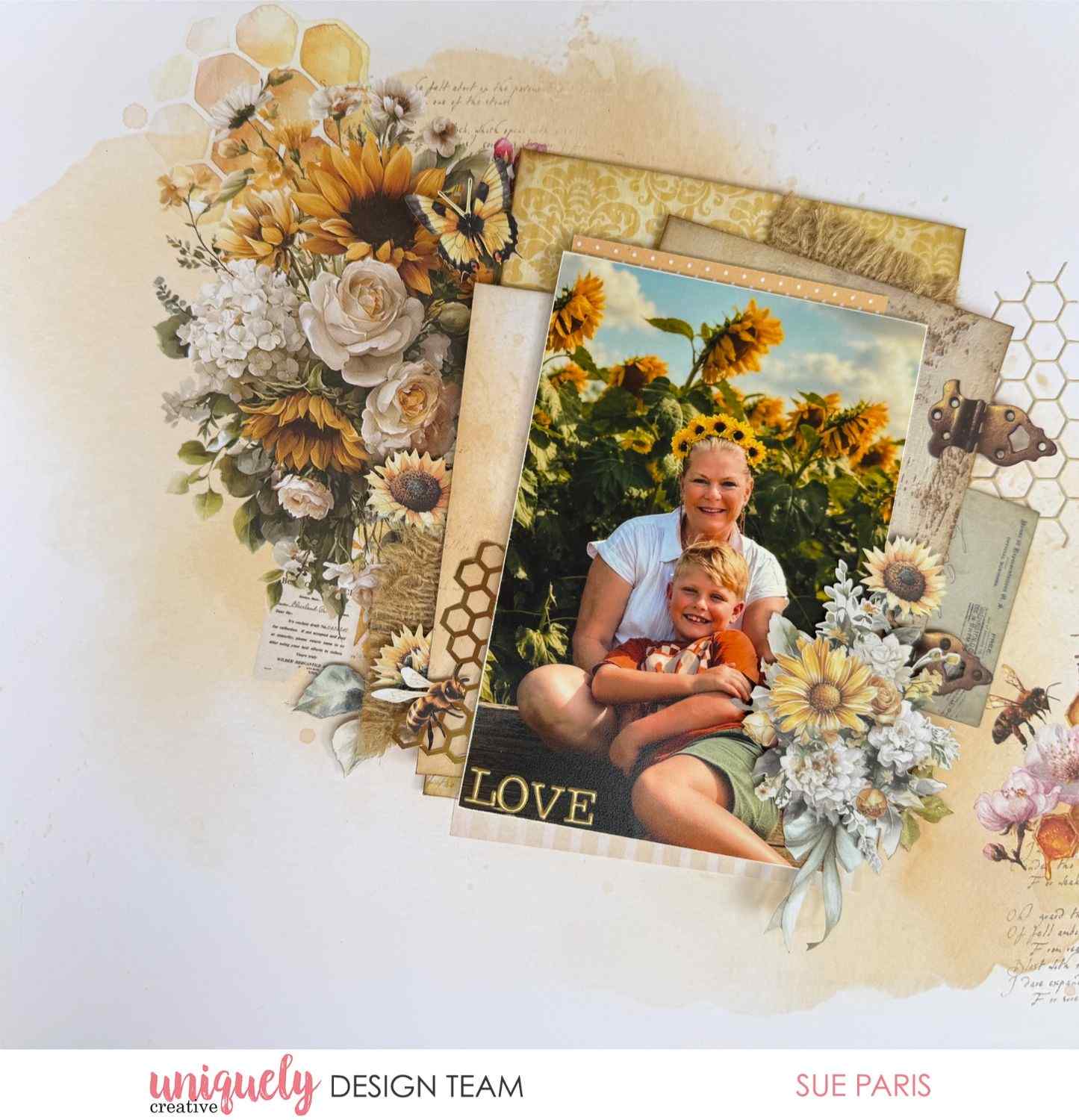 Uniquely Creative Sweet As Honey scrapbook layout with "Love" sentiment, large floral cluster of sunflowers and white roses, layered patterned papers, honeycomb die cuts, butterfly, and bees framing a photo of a woman and child in a sunflower field, created by Sue Paris