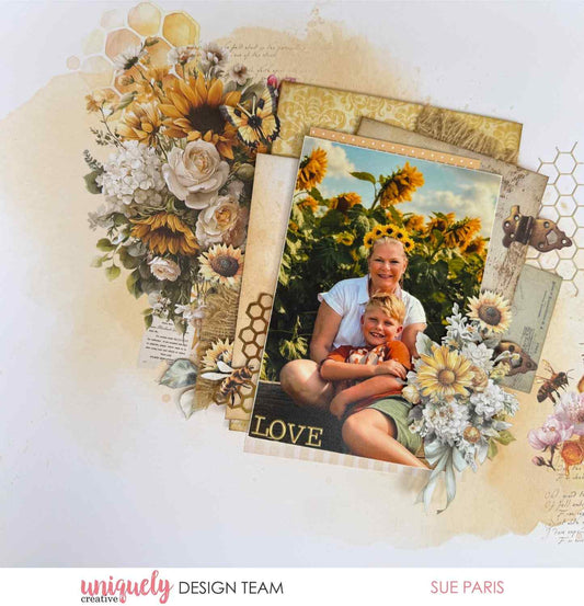 Uniquely Creative Sweet As Honey scrapbook layout with "Love" sentiment, large floral cluster of sunflowers and white roses, layered patterned papers, honeycomb die cuts, butterfly, and bees framing a photo of a woman and child in a sunflower field, created by Sue Paris