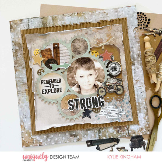 Scrapbook layout using Uniquely Creative Wings and Wheels paper, featuring boy's portrait in gear-shaped frames with Strong title, Remember to Explore sentiment, motorcycle and compass embellishments