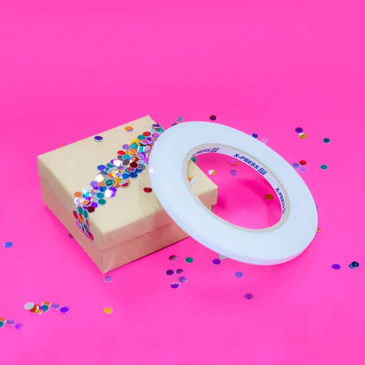 Roll of X-Press It double sided tape on a decorated gift box where they have used the tape on an angle across the box to adhere some confetti. 