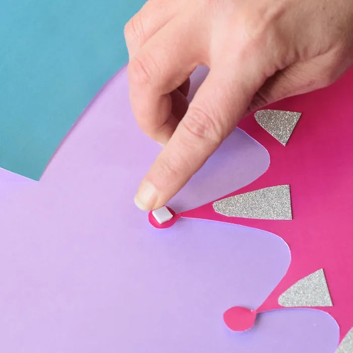 Hand placing a foam mounting square on a colourful paper craft crown. 